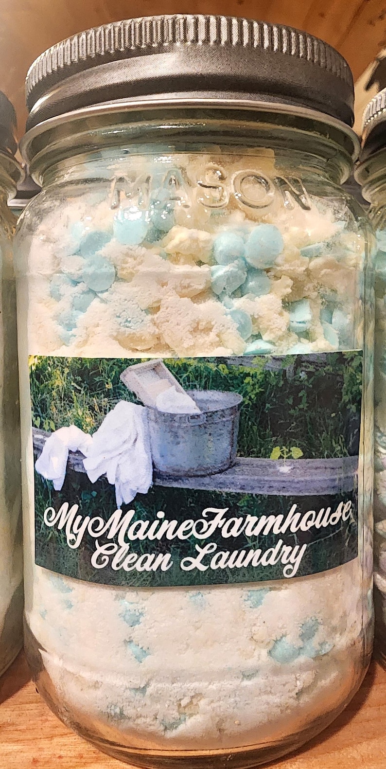 May include: A glass jar filled with white laundry detergent and blue beads. The jar has a black lid and a label that reads "My Maine Farmhouse Clean Laundry".