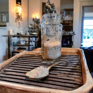 May include: A clear glass jar with a lid, filled with colorful sprinkles, sits on a brown tray with a metal spoon. The jar has the word "MASON" printed on the bottom.