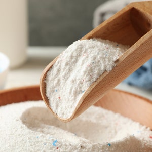 May include: A wooden scoop filled with white laundry detergent powder. The powder has small blue and red flecks throughout. The scoop is being poured into a wooden bowl filled with the same powder.