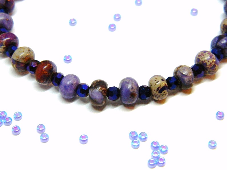 Pu&ograve; includere: Un braccialetto di perline viola e marroni con perline di vetro blu scuro. Il braccialetto &egrave; su uno sfondo bianco con perline blu iridescenti sparse.