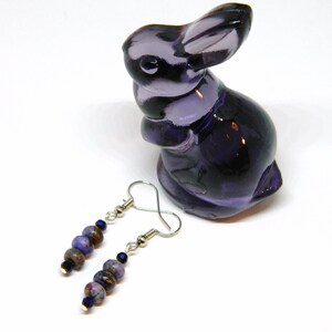 Pu&ograve; includere: Un paio di orecchini pendenti con perline viola e blu e ganci d'argento. Gli orecchini sono esposti su una superficie bianca accanto a una statuetta di coniglio di vetro viola.