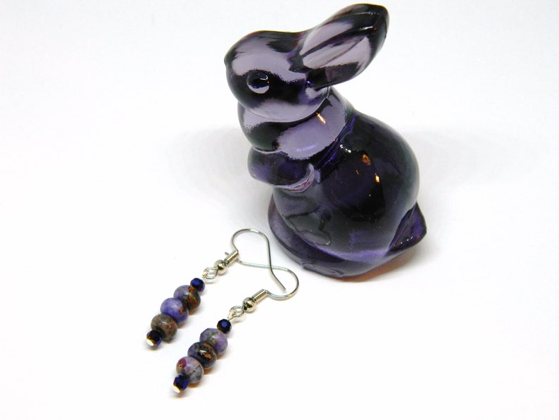 Pu&ograve; includere: Un paio di orecchini pendenti con perline viola e blu e ganci d'argento. Gli orecchini sono esposti su una superficie bianca accanto a una statuetta di coniglio di vetro viola.
