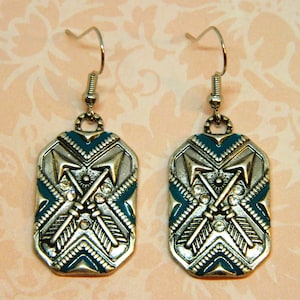 Puede incluir: Un par de pendientes de tono plateado con un diseño de esmalte turquesa y blanco. Los pendientes presentan un patrón geométrico con dos flechas cruzadas y diamantes de imitación.
