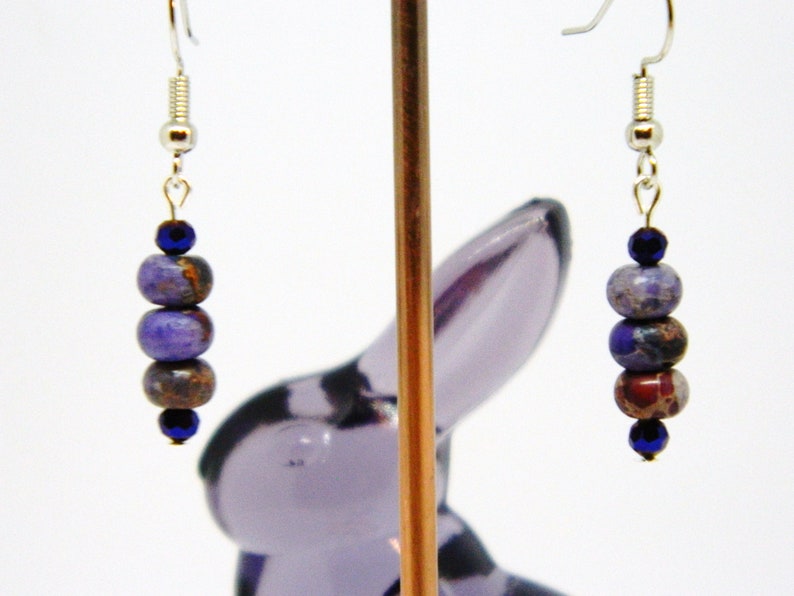 Pu&ograve; includere: Un paio di orecchini pendenti con perline viola e blu. Gli orecchini sono appesi a un gancio d'argento.