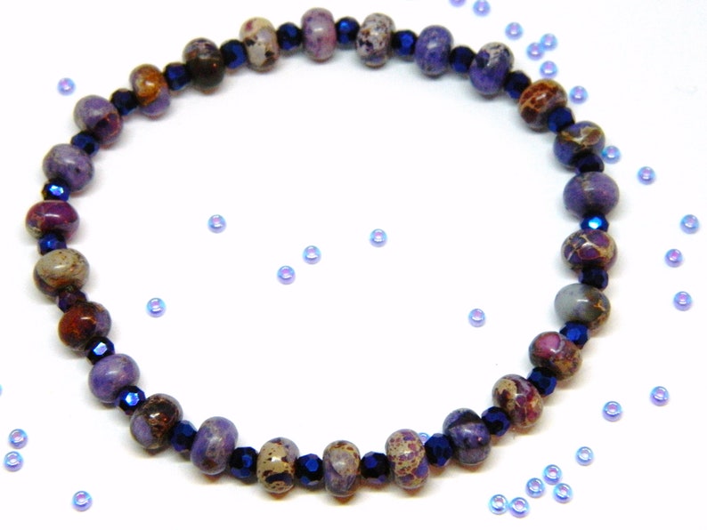 Pu&ograve; includere: Un braccialetto di pietre preziose viola e marroni con perline di vetro blu scuro. Il braccialetto &egrave; su uno sfondo bianco.