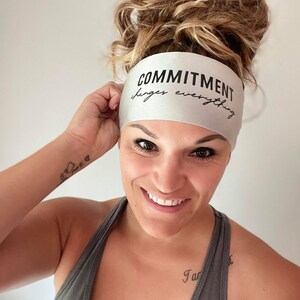 Op de afbeelding: Een vrouw draagt een zilveren hoofdband met de tekst "COMMITMENT changes everything". Ze draagt een grijs tanktop en heeft een tatoeage op haar pols.