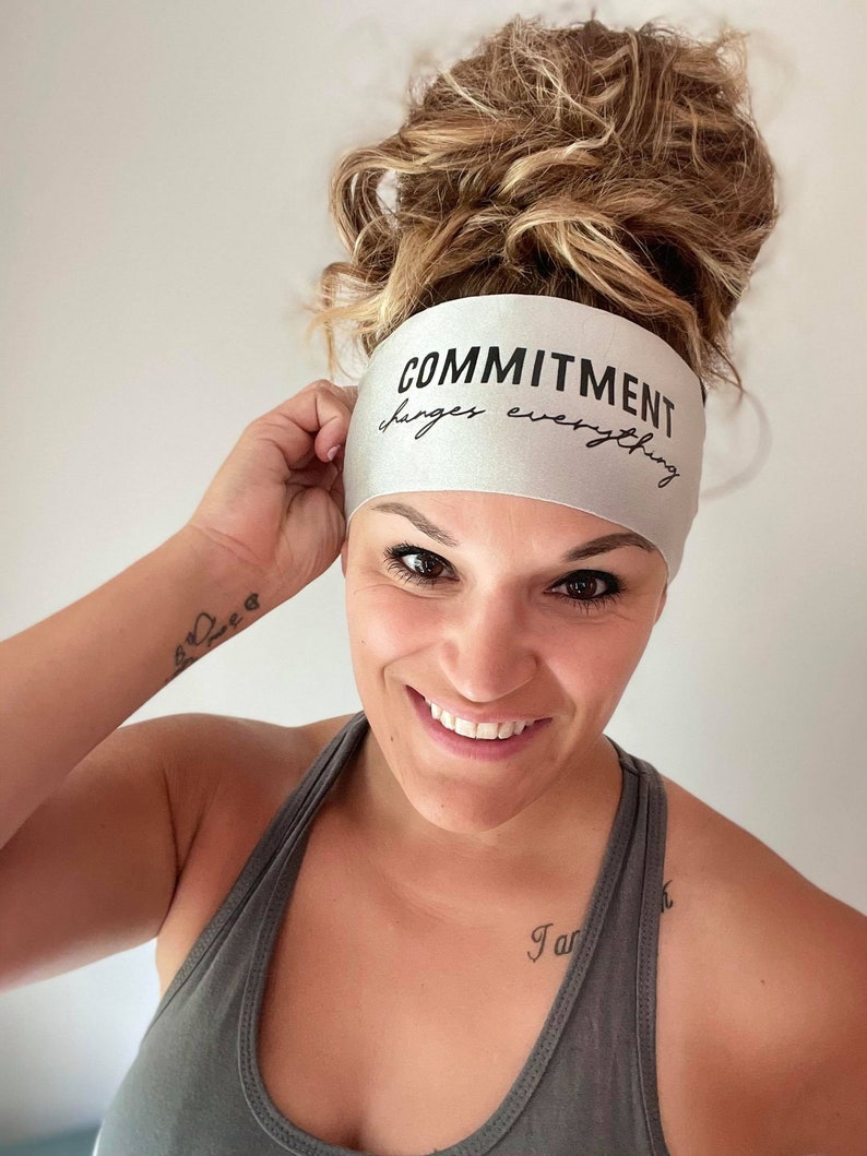 Op de afbeelding: Een vrouw draagt een zilveren hoofdband met de tekst "COMMITMENT changes everything". Ze draagt een grijs tanktop en heeft een tatoeage op haar pols.