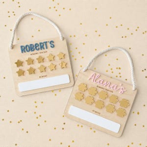 May include: Two wooden reward trackers with white rope hangers. One features the blue text ROBERT'S REWARD TRACKER with gold star tokens. The other features pink text Alana's REWARD TRACKER with gold flower tokens. Both have white rectangular labels.