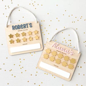 May include: Two wooden reward trackers with rope handles. One has "ROBERT'S" in blue letters and gold star-shaped tokens. The other has "Alana's" in pink letters and gold flower-shaped tokens. Both have "REWARD TRACKER" and "REWARD" text.