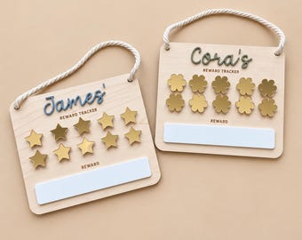 Puede incluir: Dos tableros de recompensas cuadrados de madera con colgadores de cuerda blanca. Un tablero muestra el nombre James en azul con estrellas doradas, el otro el nombre Cora en verde con flores doradas. Ambos incluyen una etiqueta rectangular blanca.