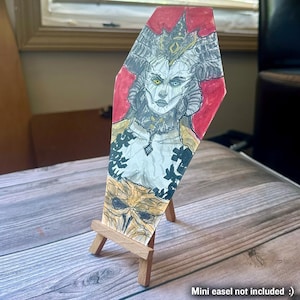 May include: A hand-drawn artwork on a coffin-shaped wooden panel. The artwork features a detailed illustration of a female figure with horns, a crown, and a stern expression. The background is red, and the figure is in shades of gray, white, and gold.
