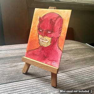 May include: A watercolor painting of a superhero in a red costume with horns, displayed on a small wooden easel. The artwork features a red and orange background. The text "Mini easel not included :)" is at the bottom.