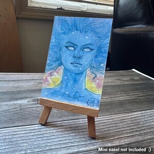 May include: A watercolor painting of a blue-skinned figure with white eyes and flowing hair, displayed on a small wooden easel. The figure has yellow and pink accents on the shoulders. The painting is signed and dated.