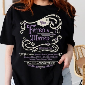 A woman wearing a black shirt that says The Fated and Mated Maasverse Tour.This is accompanied with white swirls and whirls, and other embellishments. Then it has a featuring section underneath with names from SJM books.