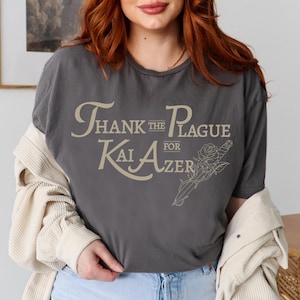 May include: A gray t-shirt with the text "Thank the Plague for Kai Aizer" in a vintage font. The text is in a light beige color and is surrounded by a floral design.