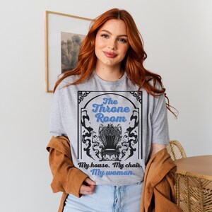 A woman wearing a sport gray colored T-shirt featuring  text that reads "The Throne Room, My House. My Chair. My Woman" accompanied by an intricate image of a throne surrounded by mysterious shadow tendrils.