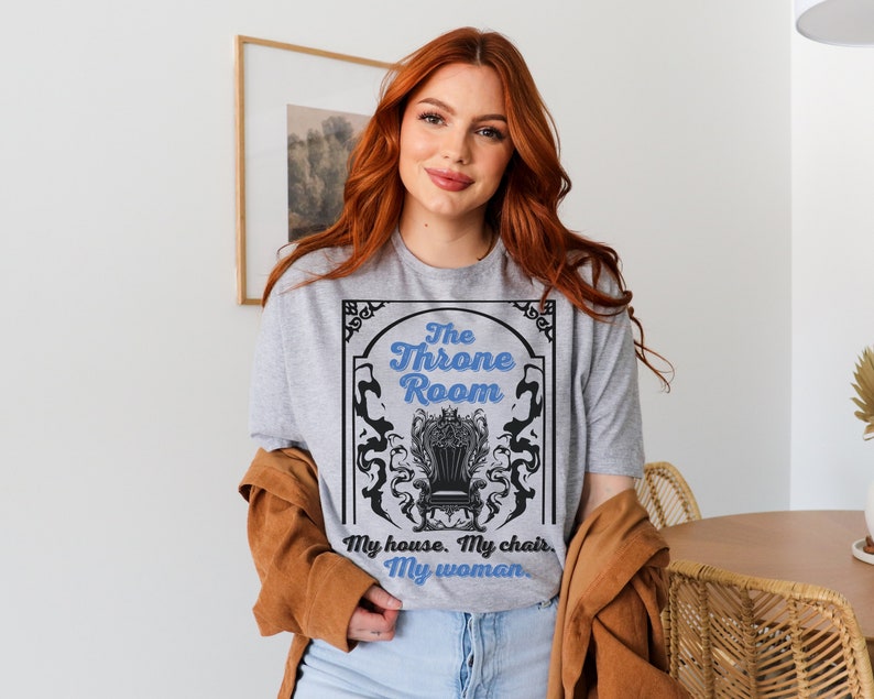 A woman wearing a sport gray colored T-shirt featuring  text that reads "The Throne Room, My House. My Chair. My Woman" accompanied by an intricate image of a throne surrounded by mysterious shadow tendrils.