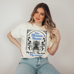 A woman wearing a white colored T-shirt featuring  text that reads "The Throne Room, My House. My Chair. My Woman" accompanied by an intricate image of a throne surrounded by mysterious shadow tendrils.