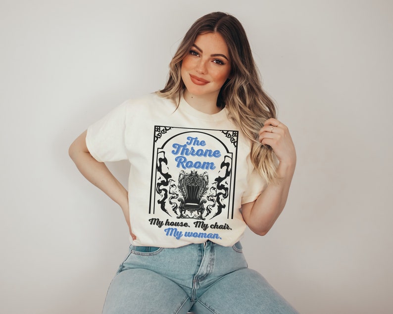 A woman wearing a white colored T-shirt featuring  text that reads "The Throne Room, My House. My Chair. My Woman" accompanied by an intricate image of a throne surrounded by mysterious shadow tendrils.