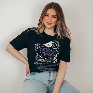 A woman wearing a black shirt that says The Fated and Mated Maasverse Tour.This is accompanied with white swirls and whirls, and other embellishments. Then it has a featuring section underneath with names from SJM books.