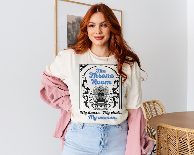 A woman wearing a natural colored T-shirt featuring  text that reads "The Throne Room, My House. My Chair. My Woman" accompanied by an intricate image of a throne surrounded by mysterious shadow tendrils.