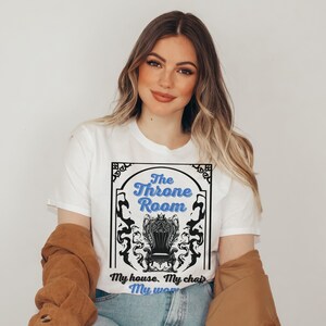 A woman wearing a white colored T-shirt featuring  text that reads "The Throne Room, My House. My Chair. My Woman" accompanied by an intricate image of a throne surrounded by mysterious shadow tendrils.