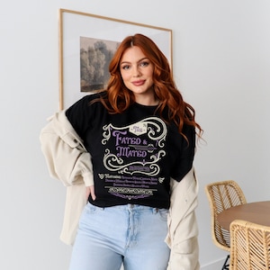 A woman wearing a black shirt that says The Fated and Mated Maasverse Tour.This is accompanied with white swirls and whirls, and other embellishments. Then it has a featuring section underneath with names from SJM books.