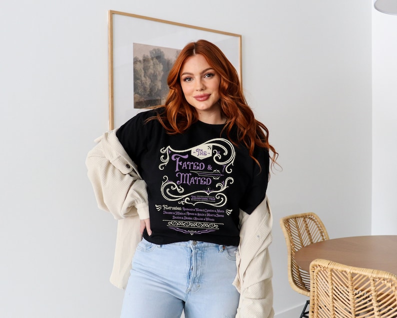 A woman wearing a black shirt that says The Fated and Mated Maasverse Tour.This is accompanied with white swirls and whirls, and other embellishments. Then it has a featuring section underneath with names from SJM books.
