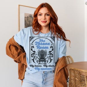 A woman wearing a light blue colored T-shirt featuring  text that reads "The Throne Room, My House. My Chair. My Woman" accompanied by an intricate image of a throne surrounded by mysterious shadow tendrils.