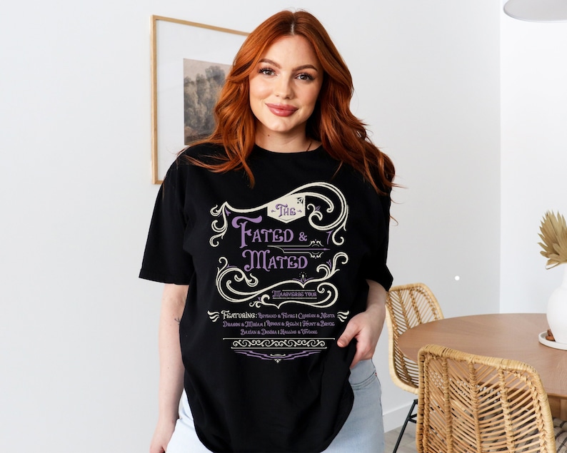 A woman wearing a black shirt that says The Fated and Mated Maasverse Tour.This is accompanied with white swirls and whirls, and other embellishments. Then it has a featuring section underneath with names from SJM books.