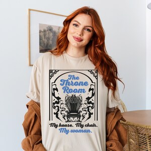 A woman wearing a sand colored T-shirt featuring  text that reads "The Throne Room, My House. My Chair. My Woman" accompanied by an intricate image of a throne surrounded by mysterious shadow tendrils.