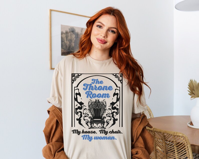 A woman wearing a sand colored T-shirt featuring  text that reads "The Throne Room, My House. My Chair. My Woman" accompanied by an intricate image of a throne surrounded by mysterious shadow tendrils.