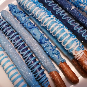 May include: A close-up of several pretzel rods dipped in chocolate and decorated with blue and white sprinkles. The pretzels are arranged on a white surface.