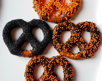 May include: Four pretzel bites covered in chocolate and sprinkles. Two pretzels are covered in black sprinkles, one in orange sprinkles, and one in black and orange sprinkles.