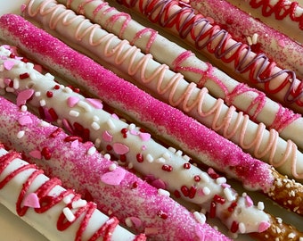 May include: A close-up of several pretzel rods dipped in white chocolate and decorated with pink and red sprinkles and drizzled with white and pink chocolate.
