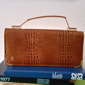 Puede incluir: Un bolso de mano marrón con estampado de cocodrilo y asa dorada. El bolso está sobre una pila de libros con el texto "Pubblicita in Italia" y los años "1977", "81 1982" y "8283" visibles.