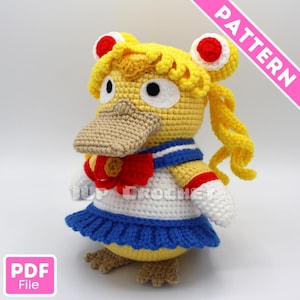 以下が含まれることがあります： ベージュのくちばしを持つ黄色いアヒルのようなキャラクターの編みぐるみ。赤色のリボンが付いた白と青のセーラー服を着ている。キャラクターの黄色い髪は2つのお団子とツインテールに結ばれている。1UP CROCHETとPATTERNの文字が記載されている。
