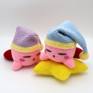 Pu&ograve; includere: Due peluche rosa all'uncinetto con gli occhi chiusi e cappelli gialli e viola stanno dormendo su una stella gialla all'uncinetto.