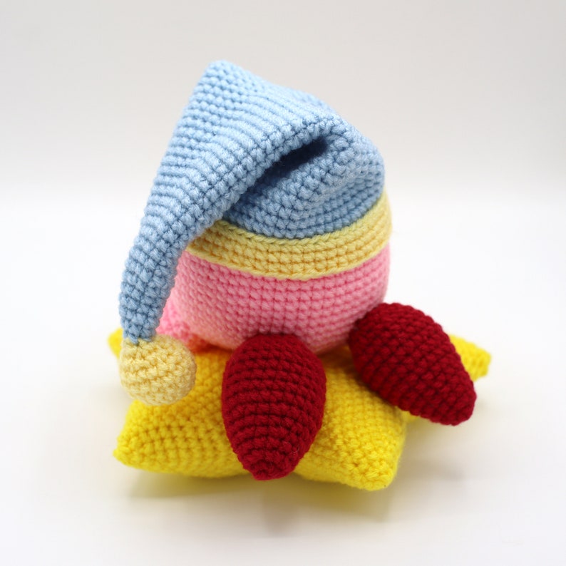 Pu&ograve; includere: Un giocattolo razzo all'uncinetto con un berretto da notte blu, un corpo rosa e giallo e fiamme rosse. Il razzo &egrave; seduto su una stella gialla all'uncinetto.