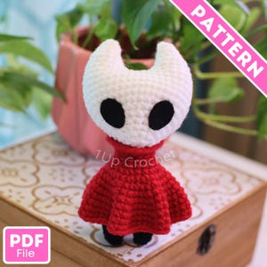 Peut inclure: Une figurine crochetée faite à la main, avec une tête blanche, des yeux noirs et une robe rouge. La figurine est posée sur une surface en bois. Le texte "1Up Crochet" est visible. Une bannière rose avec le mot "PATTERN" est en arrière-plan.