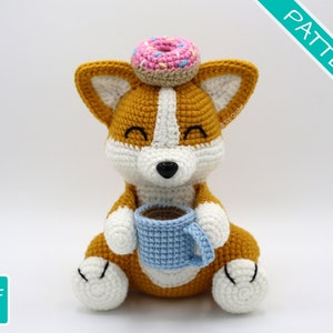 Könnte beinhalten: Ein gehäkeltes Amigurumi-Corgi, das einen rosafarbenen und weißen Donut auf dem Kopf trägt und eine blaue Kaffeetasse hält.