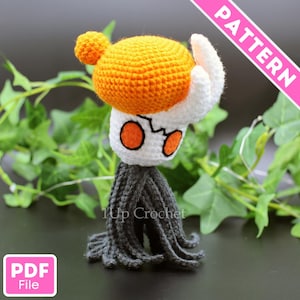 Può includere: Una figura all'uncinetto fatta a mano con una testa bianca, occhi arancioni e una grande testa arancione. La figura ha una base grigia con frange ed è ambientata su un fogliame verde. La parola "PATTERN" è visibile.