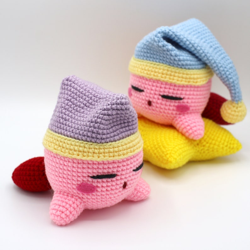 Pu&ograve; includere: Due peluche rosa all'uncinetto con gli occhi chiusi e cappucci da notte rossi e viola sono sdraiati su una stella gialla all'uncinetto.