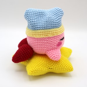 Pu&ograve; includere: Un giocattolo di peluche rosa all'uncinetto con un cappello blu e un bordo giallo, seduto su una stella gialla all'uncinetto.