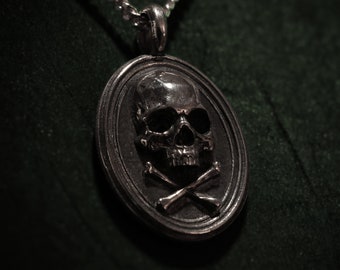 May include: A silver pendant necklace with a skull and crossbones design inside an oval frame. The skull has detailed features, and the crossbones are beneath. The pendant hangs from a silver chain, set against a dark green backdrop.