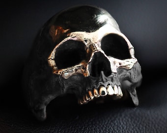 May include: A silver skull ring with a detailed design. The skull has a prominent brow, deep eye sockets, and a defined jawline. The teeth are visible and slightly open.