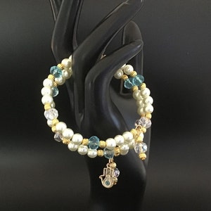 Puede incluir: Un brazalete de doble cadena con perlas blancas, cuentas doradas y cuentas de cristal azul. Un dije dorado con un ojo azul cuelga del brazalete.