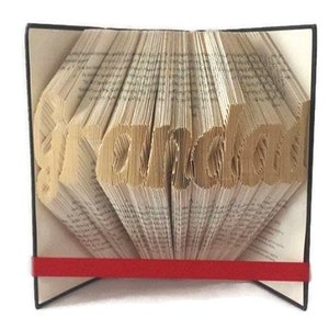 May include: A book art sculpture with the word "remarkable" folded into the pages. The book is bound in black with a red ribbon.