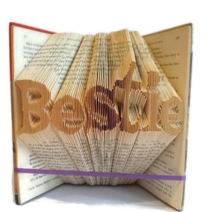 Könnte beinhalten: Ein Buch mit dem Wort "Bestie" in die Seiten gefaltet. Das Buch wird mit einem lila Gummiband zusammengehalten.