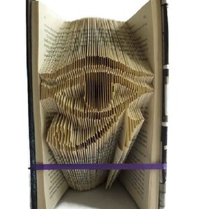 May include: A book with pages folded into a three-dimensional eye shape. The book is bound in black fabric with white stripes and is held closed with a purple elastic band.
